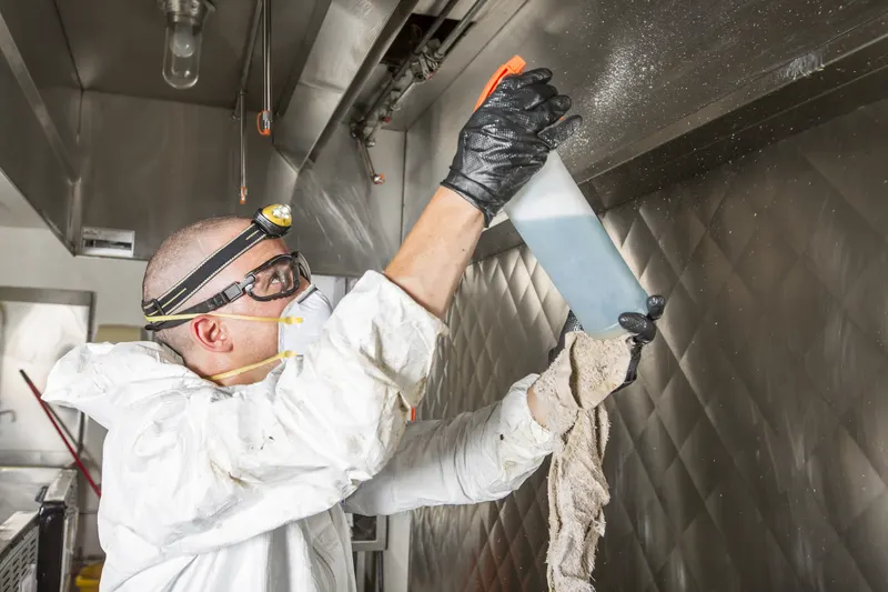 Technician in white coveralls, goggles and a headlamp wiping the underside of a stainless steel exhaust canopy with a spray bottle and cloth, seen from below