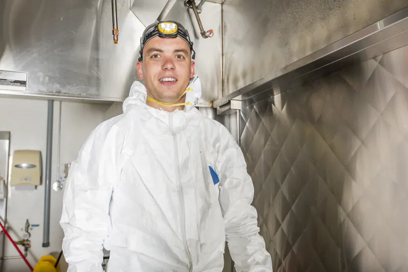 Technician in coveralls and a respirator standing beside a stainless steel cook line under a commercial kitchen hood