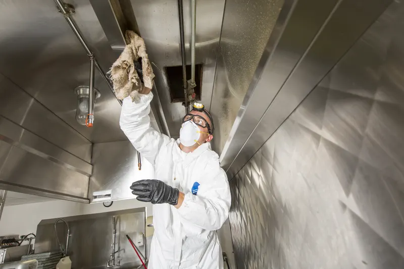 Technician in coveralls wiping grease from the plenum above the filter bank inside a stainless steel exhaust hood