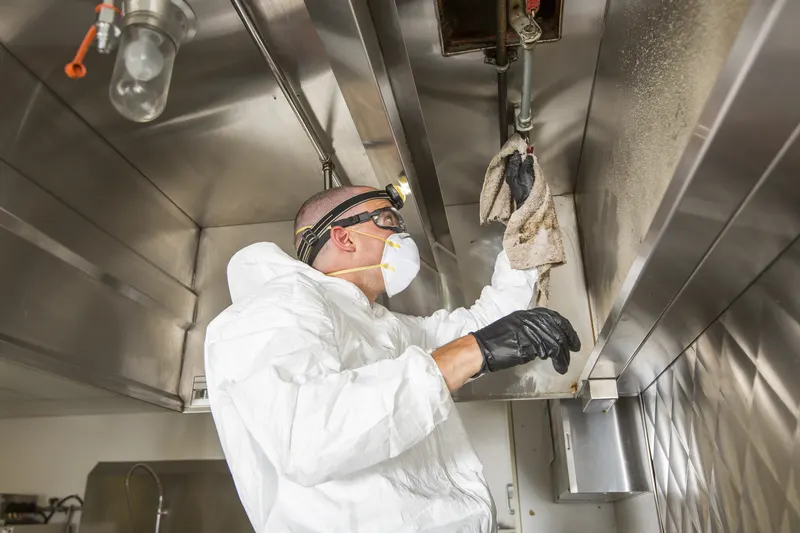 Gloved hand wiping grease from the rolled front edge of a stainless steel kitchen hood