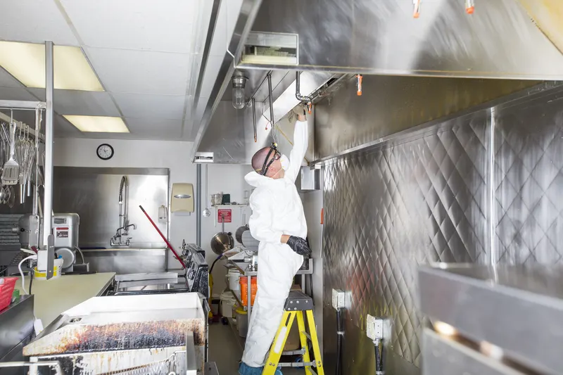 Technician on a step ladder wiping down a stainless steel exhaust canopy above a covered cooking line